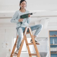 young woman in her new flat sitting on ladder, holding electric drill - home decoration stockfoto's en -beelden