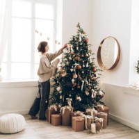 young woman is decorating christmas tree with many different decorations and festive garland in minimalist light interior. new year celebration concept - home decoration stockfoto's en -beelden