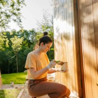young woman painting an exterior wall of a wooden house - home decoration stock pictures, royalty-free photos & images