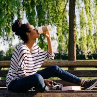 young woman sitting on park bench drinking coffee to go - junk food stock pictures, royalty-free photos & images