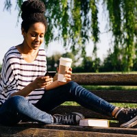 young woman sitting on park bench with a book and coffee to go looking at smartphone - junk food stock pictures, royalty-free photos & images