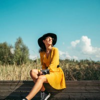 young woman wearing a black hat and yellow dress with an analog camera sitting on wooden boardwalk - fashion photos et images de collection
