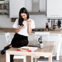 young woman with cell phone eating pizza in kitchen at home - junk food stock pictures, royalty-free photos & images