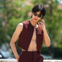 Yugo Takano wears brown sunglasses, silver large chain long necklaces, a brown burgundy sleeveless / cropped gilet, matching brown burgundy flared...