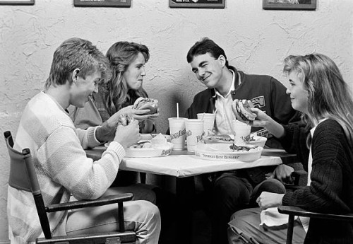1980s GROUP OF FOUR TEENS SITTING AROUND TABLE AT RESTAURANT CHATTING & EATING HAMBURGERS
