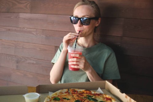 a beautiful stylish girl in sunglasses drinks a soft drink or cocktail through a straw, outdoors in a cafe. italian cuisine. sliced pizza in paper packaging on a wooden table. delicious lunch, snack, fast food. - junk food st