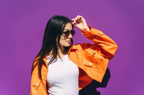 a beautiful young asian woman in sunglasses and a bright orange jacket on a purple background. - fashion stock pictures, royalty-free photos & images