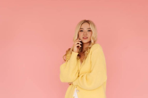 a beautiful young blonde woman with curly hair in a yellow knitted cardigan is chatting on the phone, communicating using a mobile phone while standing on a pink isolated background. place for text - fashion stock pictures, r