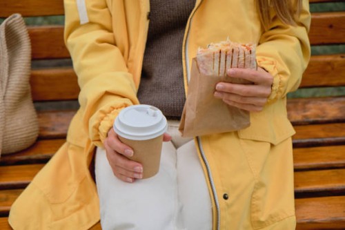 a beautiful young woman or girl holds a disposable mug of coffee in her hands, drinks it, eats a delicious vegetarian or vegan shawarma doner kebab while sitting on a park bench. the concept of fast food and unhealthy food, s