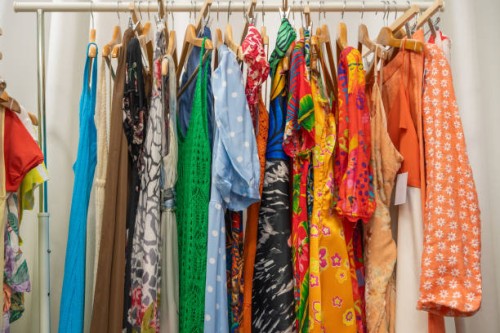 a colorful selection of vibrant dresses and garments hanging on a clothing rack, featuring an array of bold patterns and bright hues, from polka dots and floral prints to abstract designs from a second-hand vintage shop. spai