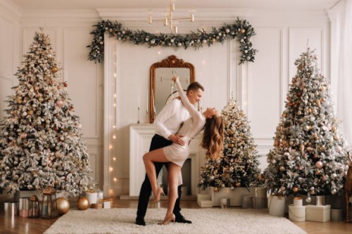 a couple in love in elegant fashionable outfits embrace, rejoice and celebrate the christmas holiday in a bright luxury decorated interior with christmas trees in the living room of the house. a young man in a suit and a wom