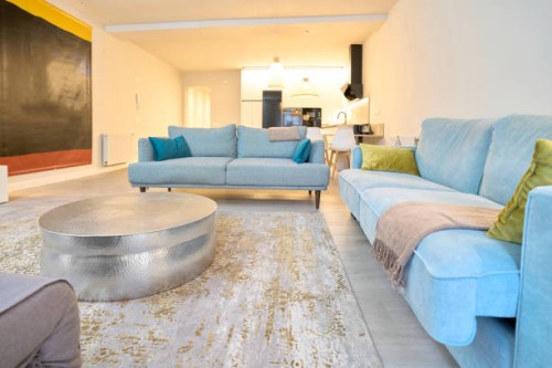 a living room set, light blue furniture and blue cushions, a round silver mousetrap table, a tv and a picture on the side, in the background you can see the kitchen and two lit lamps, an exit sign from the apartment. - home d