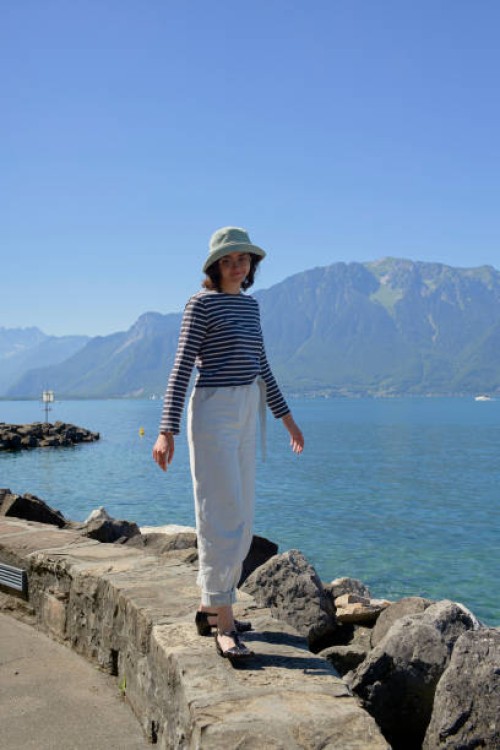 a person stands on a rocky shoreline with a large body of water and mountains in the background, wearing a hat, striped top, and white pants - fashion stock pictures, royalty-free photos & images
