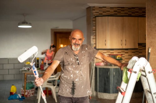 a senior wall painter poses in portrait with tools in hand smiling and looking at camera, full of paint with a roller - home decoration stock pictures, royalty-free photos & images