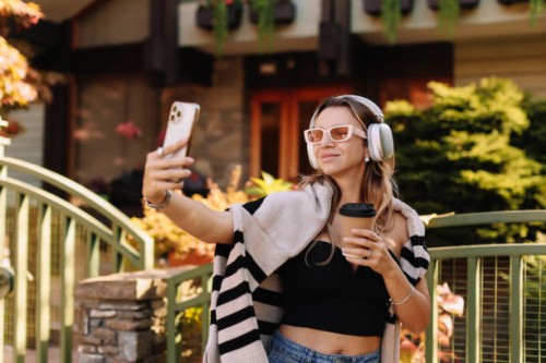 a smiling woman in sunglasses and wireless headphones takes selfies using a mobile phone and listens to music while walking along an autumn street in the countryside outdoors - fashion stock pictures, royalty-free photos & im
