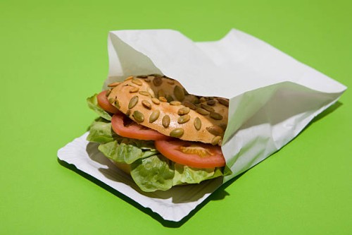a sunflower seed bagel sandwich with tomato and lettuce in a paper bag - junk food stock pictures, royalty-free photos & images