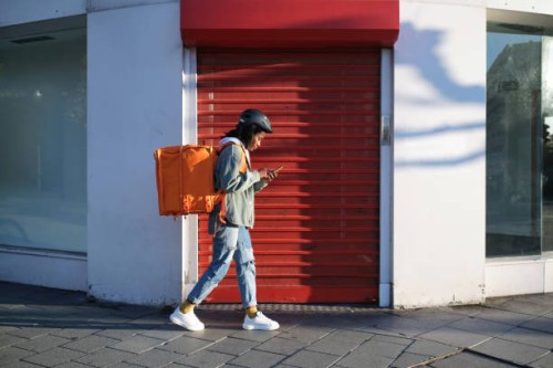 a young delivery woman with her backpack using her phone and walking in search of the delivery place - food stock pictures, royalty-free photos & images