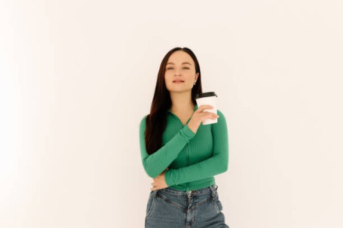a young woman in a green sweatshirt holds a glass of coffee and smiles at the camera while standing against a white isolated background. space for the text. a place for advertising. copy space - junk food stock pictures, roya