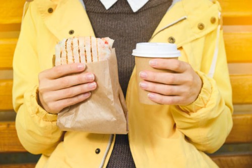 a young woman or girl holding a cup of coffee, drinking it, eating a delicious shawarma doner kebab, sitting on a bench in a public park. fast food and snack concept. - junk food stock pictures, royalty-free photos & images