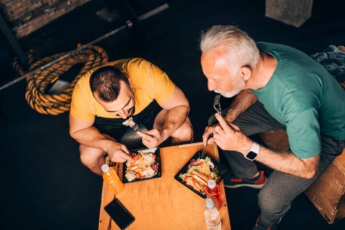 active senior sportsman eating a healthy protein salad meal in a take-out box with his personal trainer in a gym area of a gym - junk food stock pictures, royalty-free photos & images