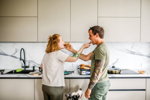 adorable couple enjoys cooking together in their well-lit, modern kitchen, playfully sharing bites of food. - food stock pictures, royalty-free photos & images