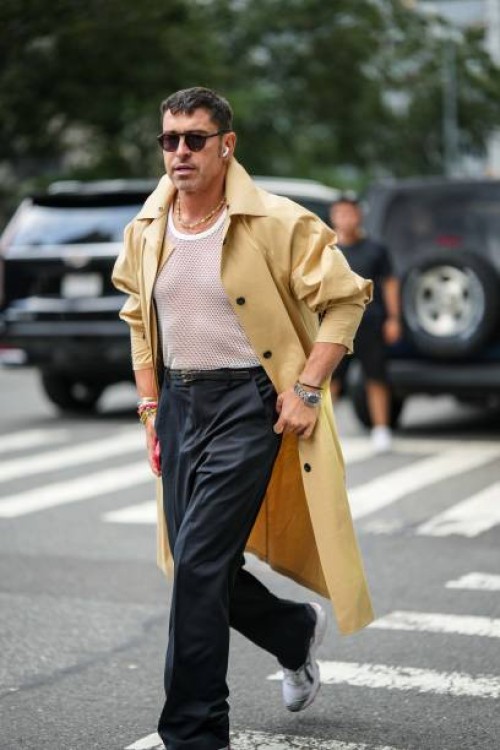 Alex Badia wears airpods Apple, a beige trench coat, a necklace, a white mesh top, a belt, black pants, Asics sneakers, outside Proenza Schouler,...