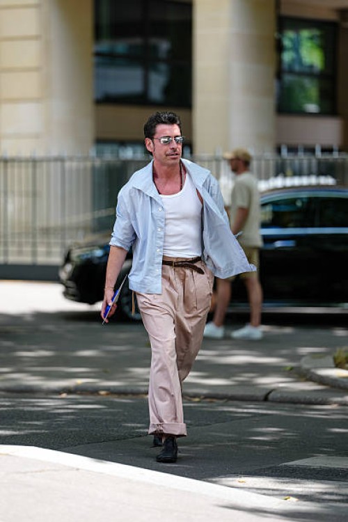 Alex Badia wears silver metallic sunglasses, dark brown beaded necklaces, white tank top, light blue unbuttoned long sleeve shirt, shiny dark brown...