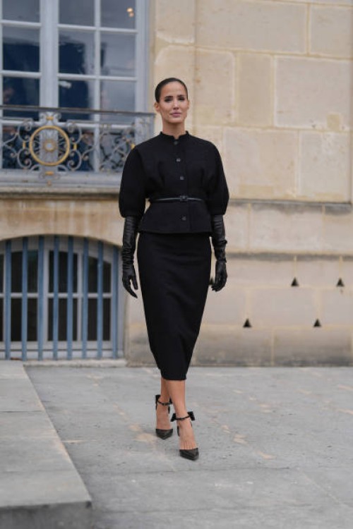 Alex Riviere wears black puffy short sleeve tailored Dior blazer jacket, black leather belt, black Dior midi skirt, shiny black leather gloves, black...