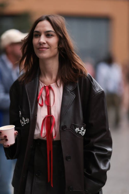 Alexa Chung is seen wearing a knee-length black leather jacket with silver gemstone details, underneath a rose top with red ribbons and a black...