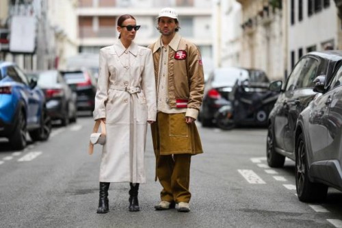 Alice Barbier wears black sunglasses, a beige suede shoulder-pads / belted long coat, a beige and white shiny leather handbag, black shiny leather...