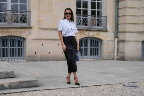 Amina Muaddi wears black sunglasses, silver earrings, white buttoned up short sleeve shirt, black high waisted midi skirt, black pointed toe leather...