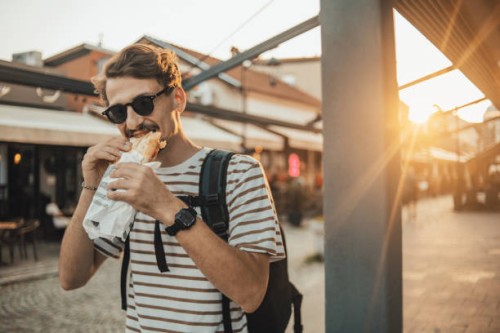 an adult fashionable man is walking in the city and eating a sandwich - junk food stock pictures, royalty-free photos & images
