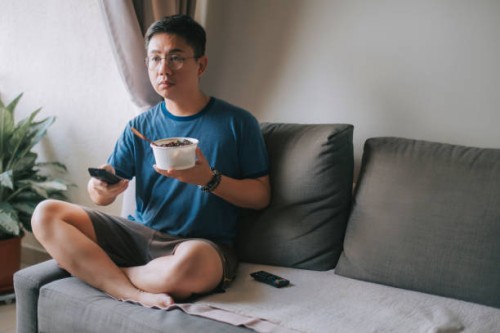 an asian chinese mid adult man having his take out food while pressing tv remote control watching tv on sofa - junk food stock pictures, royalty-free photos & images