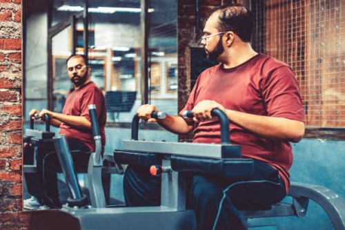 an overweight man at a gym using exercising machine while looking at the mirror - junk food stock pictures, royalty-free photos & images