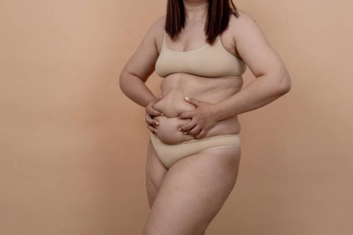 an unrecognizable woman in beige underwear clutches the folds of a thick belly with her hands on a beige isolated background - junk food stock pictures, royalty-free photos & images