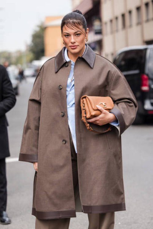 Ashley Graham, wearing a brown trench coat and trousers, a light blue and white striped shirt and and light brown leather bag, is seen outside Tod's...