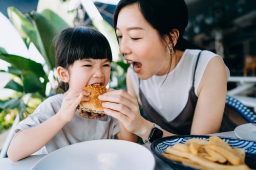 asian family having fun while enjoying lunch in an outdoor restaurant, mother and daughter are sharing and having a big bite eating a burger. family eating out lifestyle - junk food stock pictures, royalty-free photos & image