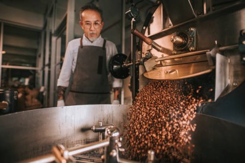asian senior man craftsperson observing freshly roasted coffee beans being removed from the roaster into the cooling cylinder with motion blur - coffee stock pictures, royalty-free photos & images