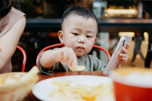asian toddler boy eating french fries and playing on smartphone in a restaurant - junk food stock pictures, royalty-free photos & images