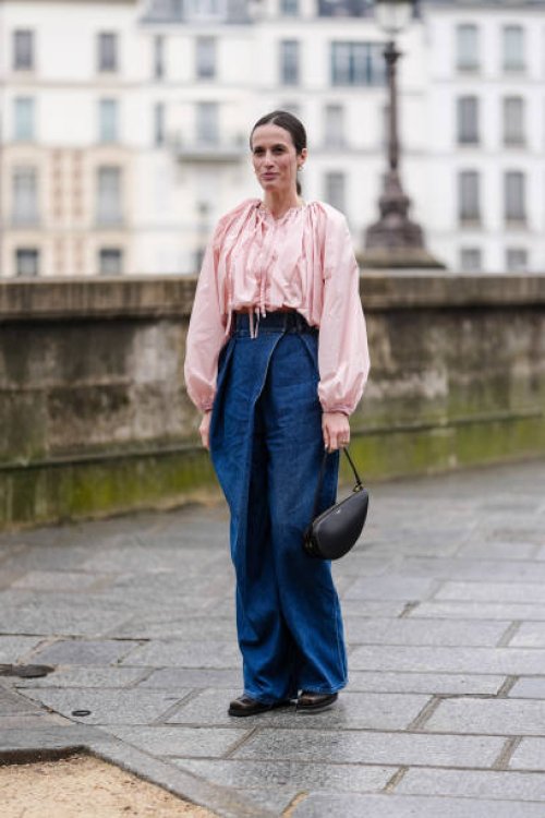 Astrid Boutrot wears gold earrings, light pink long sleeve shirt, navy blue high waisted denim jean pants, black Alaïa leather bag, shiny dark brown...