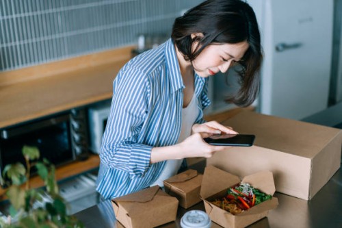 beautiful and cheerful young asian woman standing by the kitchen counter, taking a photo of delicious home delivery takeaway fresh healthy salad lunch box with smartphone, sharing on social media before eating it. eating at h