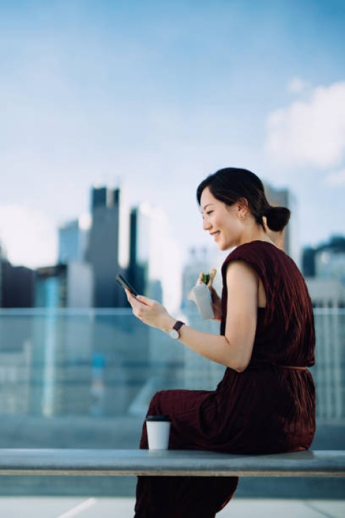 beautiful smiling young asian businesswoman sitting on the bench in office building terrace, against corporate skyscrapers. using smartphone outdoors while eating a healthy sandwich during lunch break. lifestyle and technolog