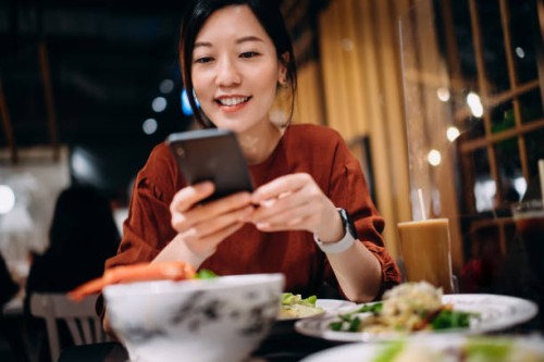 beautiful smiling young asian woman taking photos of scrumptious traditional thai food served on dining table with smartphone while enjoying lunch in a thai restaurant. eating out lifestyle. camera eats first culture - food s