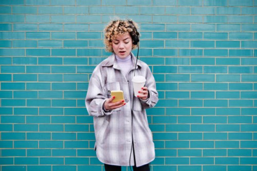 beautiful woman with coffee cup listening music through headphones while using smart phone against blue wall - junk food stock pictures, royalty-free photos & images