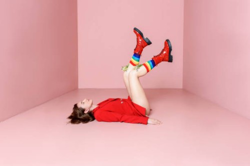 beautiful young woman in a red jacket posing while lying on a pink background in colored rainbow socks from which flowers grow and boots. fashion concept. - fashion stock pictures, royalty-free photos & images