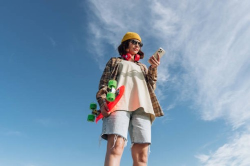 beautiful young woman in summer clothes using smart phone and holding skateboard on blue sky background. internet concept - fashion stock pictures, royalty-free photos & images