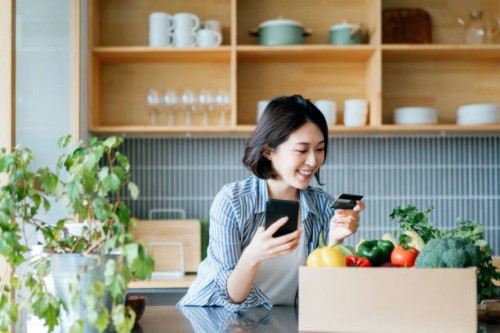 belle jeune femme asiatique souriante faisant ses courses en ligne avec l’appareil d’application mobile sur le smartphone et faisant le paiement en ligne avec sa carte de crédit, avec une boîte d’épicerie organique colorée et