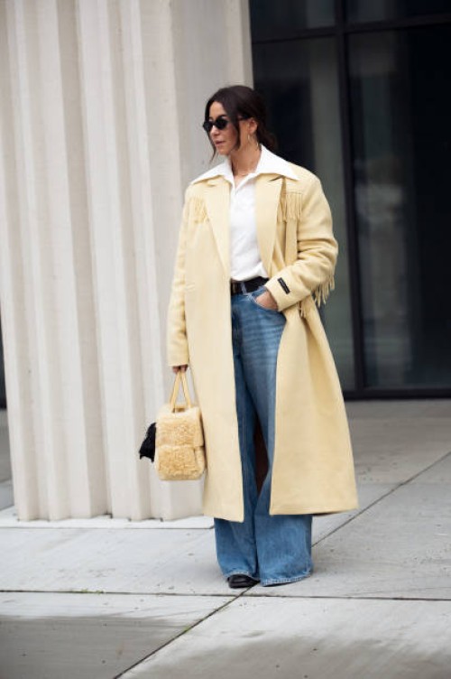 Benthe Liem wears blue jeans, white shirt, pale yellow long coat, yellow bag with attached black mini bag outside the Herskind fashion show during...