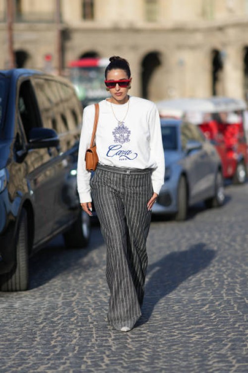 Bettina Looney wears red sunglasses, a white pullover, a brown leather bag, gray striped pants, outside Casablanca, during the Womenswear...