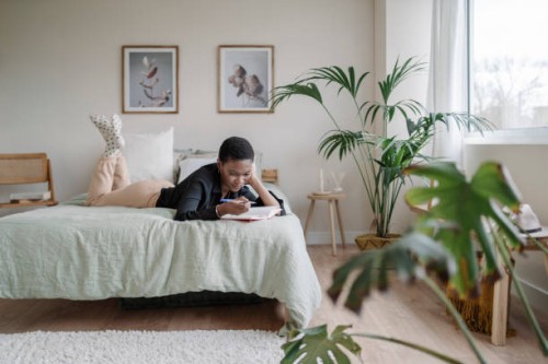 black woman writing in a notebook while laying on a comfortable bed at home. - home decoration stock pictures, royalty-free photos & images